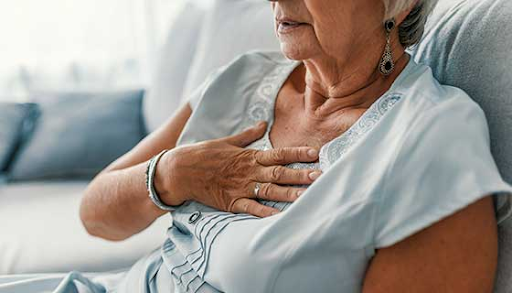 Elderly woman sitting on a couch, holding her chest due to breathing difficulty.