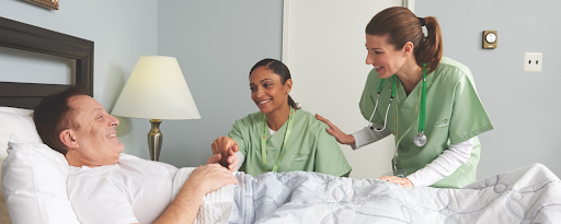 A smiling patient in his home bed interacting with two nurses in green uniforms, symbolizing compassionate care.